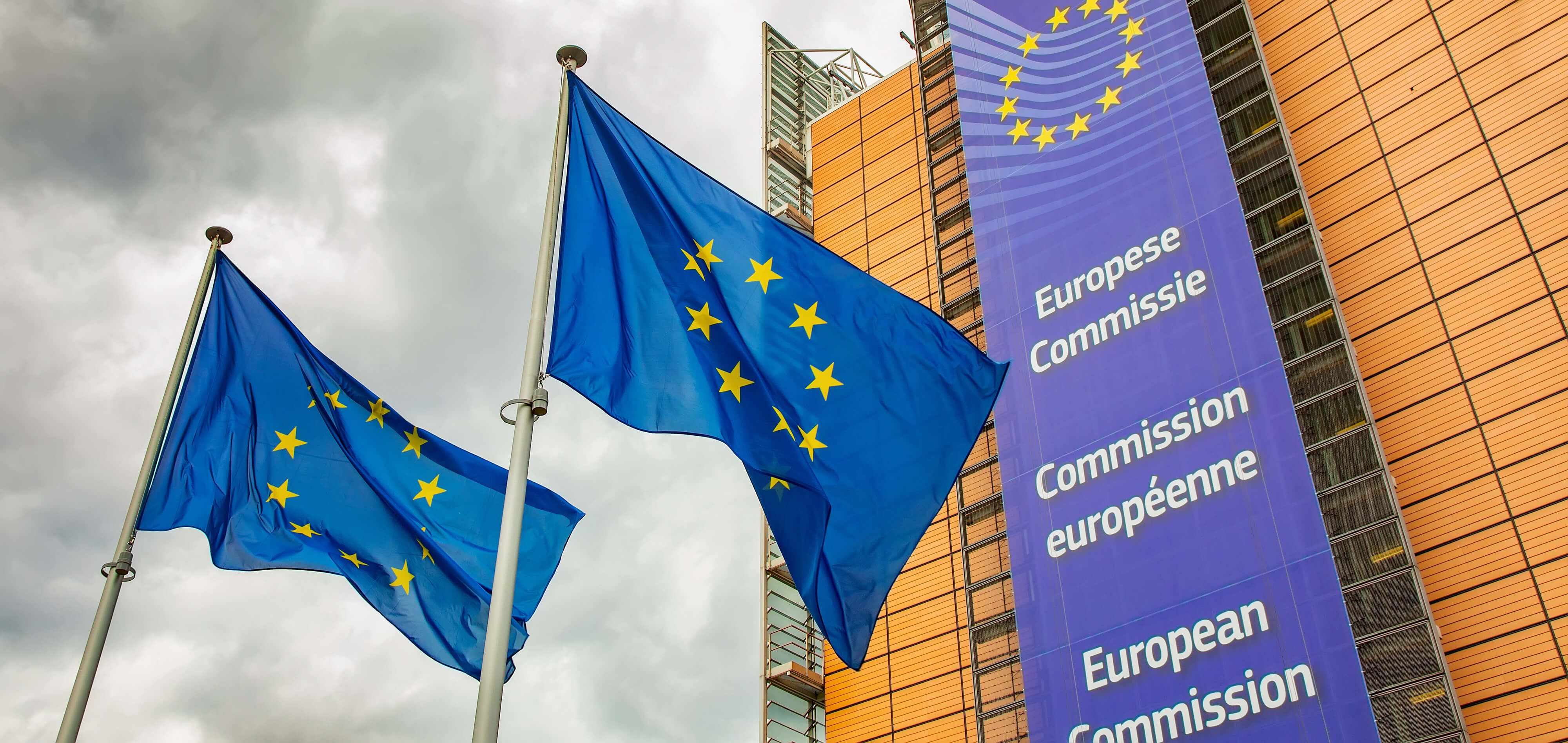 EU flags in front of the European Commission building with an overcast sky in the background
