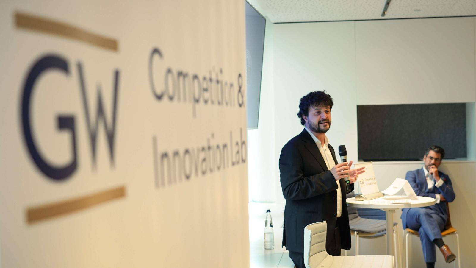 a man in a suit gives a conference presentation beside a GW sign