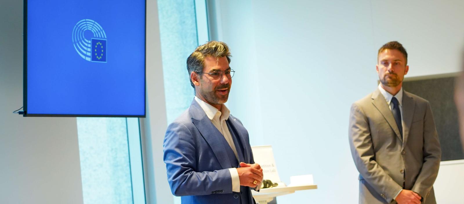 Two men - one in a blue suit and the other in a tan suit - stand at the front of a classroom giving a lecture